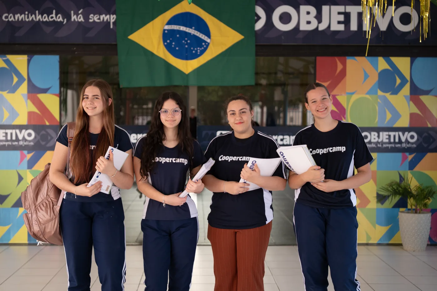 Quatro alunas de uniforme sorrindo com cadernos na entrada da escola